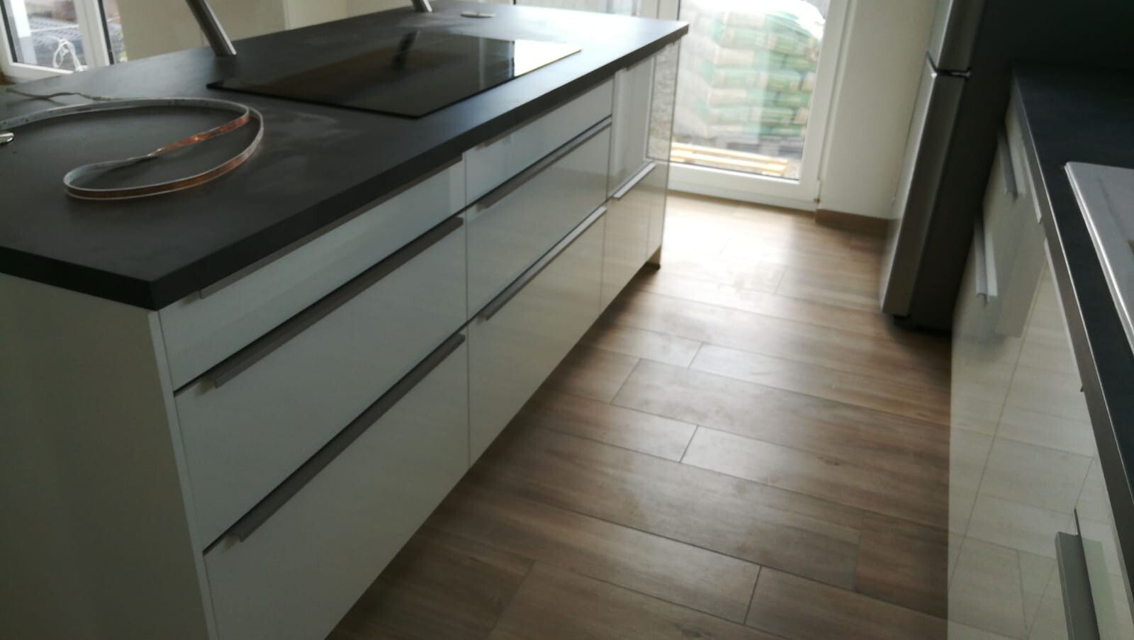 Moderne Küche mit Insel, weißen Schubladen, schwarzer Arbeitsplatte, Holzboden und Glastür am Ende.