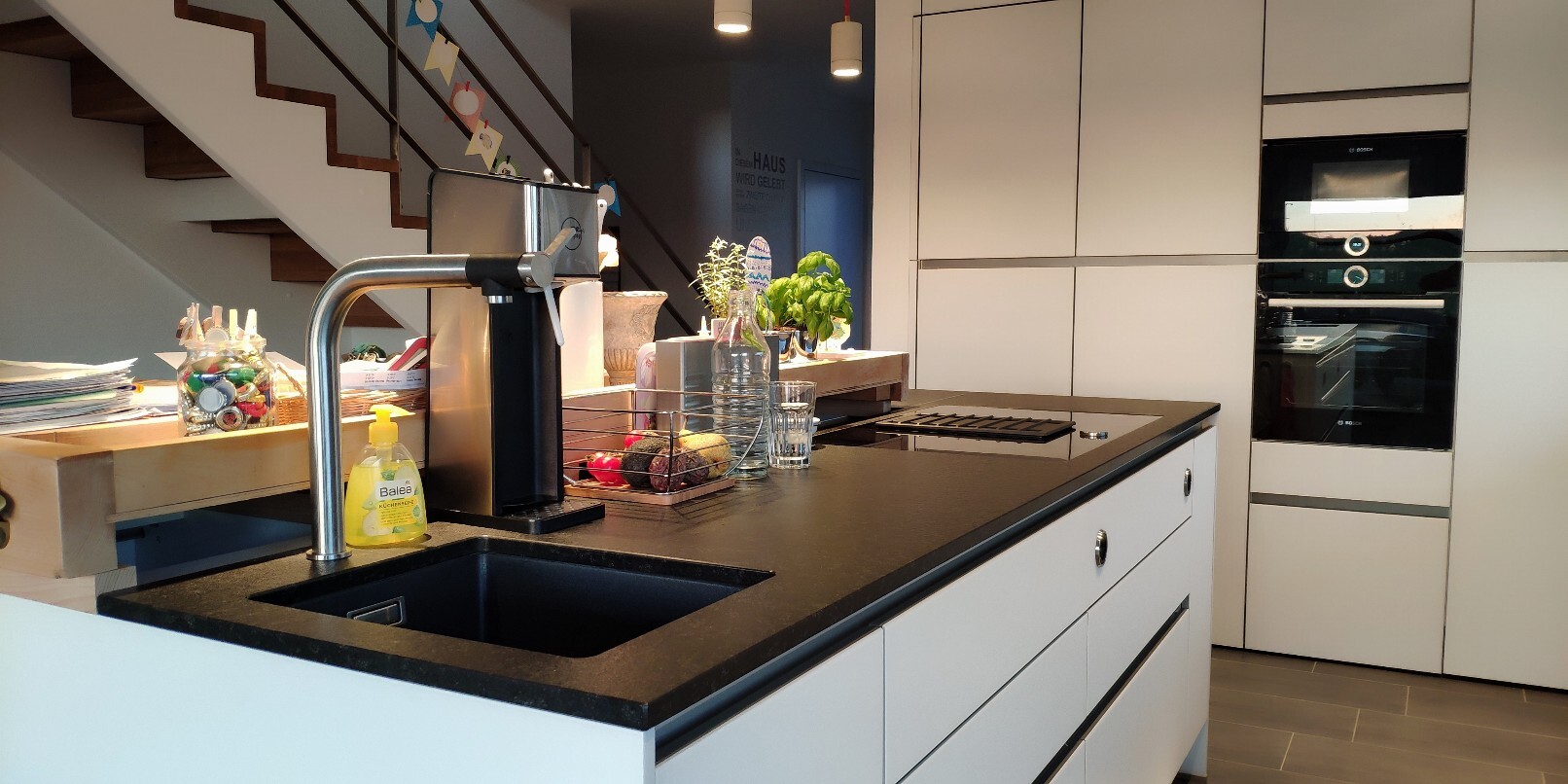 Moderne Küche mit Insel, Spüle und Wasserhahn, Obst und Kräuterpflanzen, integrierter Backofen.