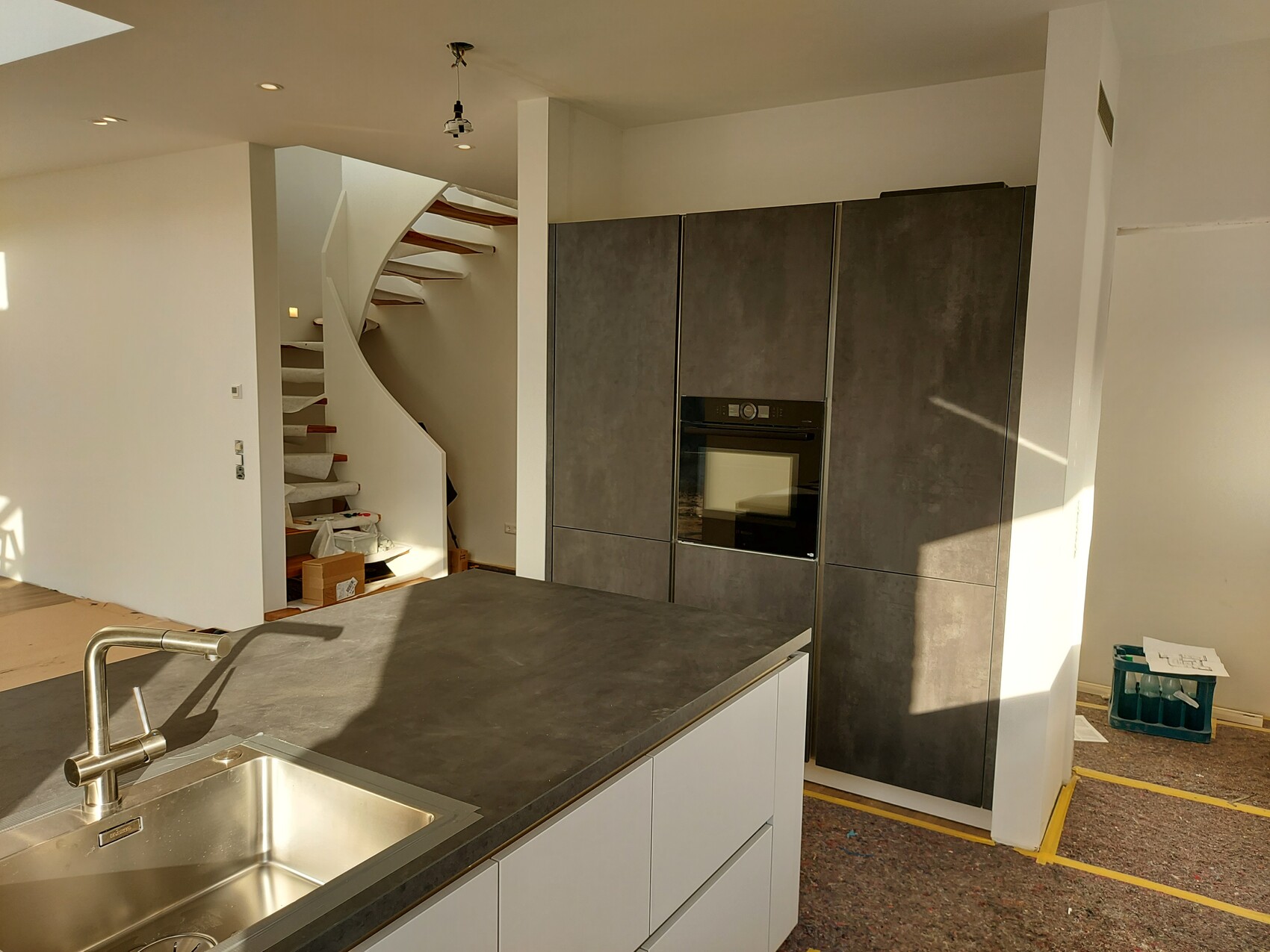 Moderne Küche mit dunkler Insel-Arbeitsplatte, Spüle, Ofen; weiße Wendeltreppe im Hintergrund.