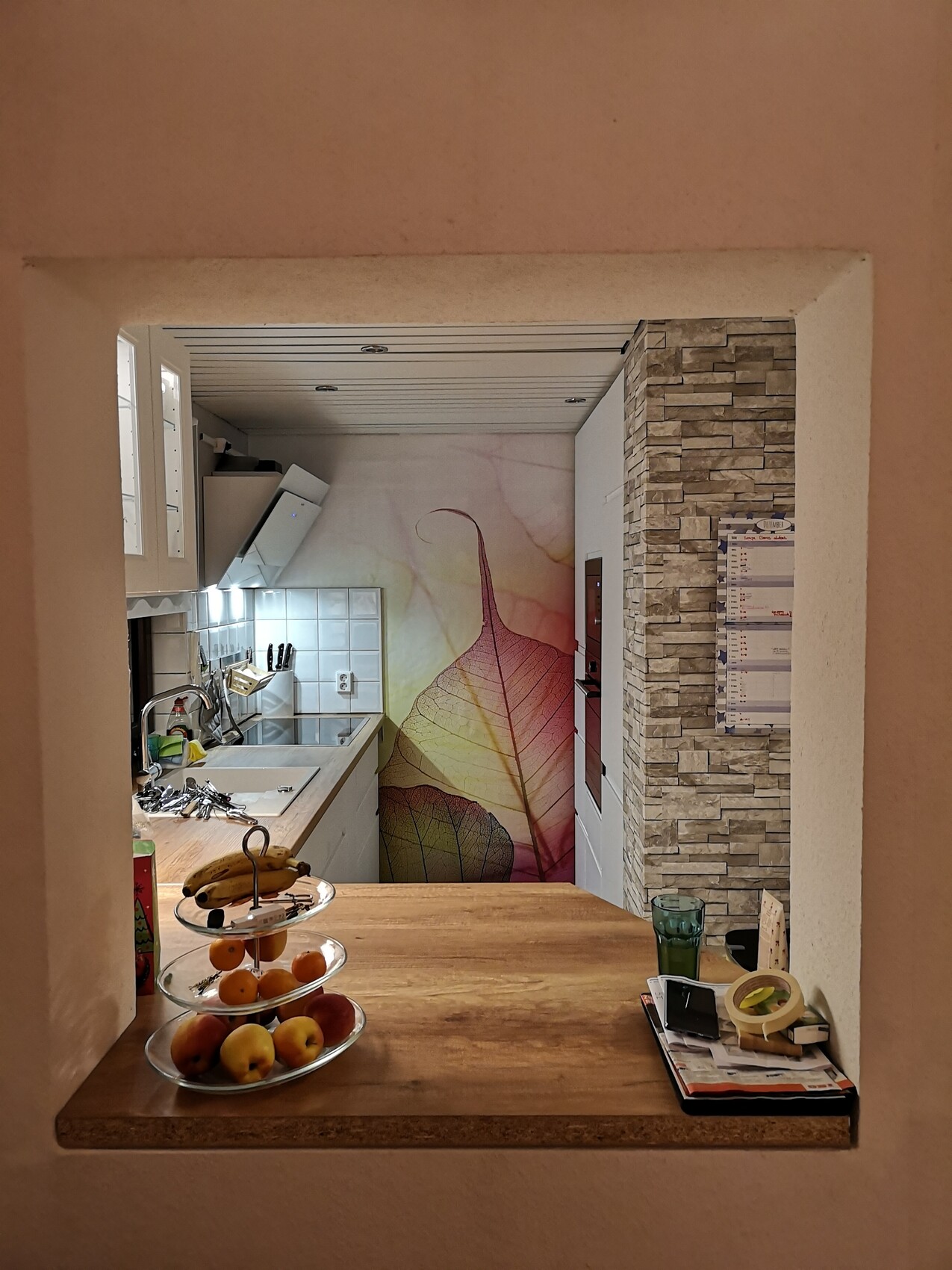 Küche hinter Wandöffnung: Obstkorb auf Holztheke, Smartphone, Kalender, bunte Blatt-Wand.