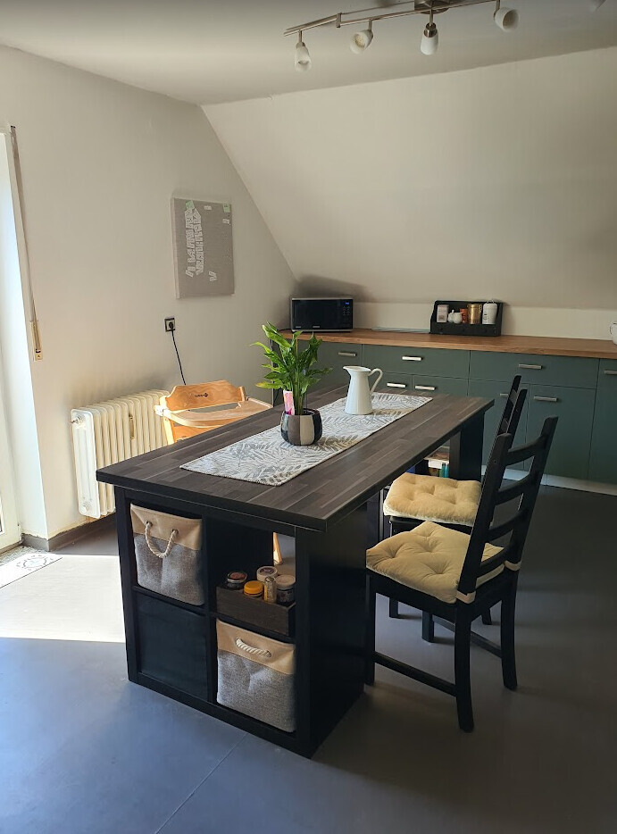 Moderne Küche mit dunkler Tischinsel, Körben darunter, Pflanze und Küchenzeile im Hintergrund.