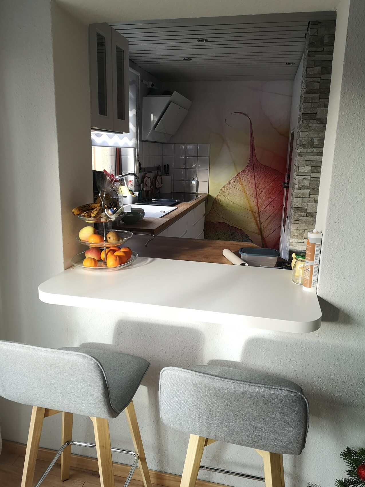 Offene Küche mit Bar-Theke, zwei graue Hocker, Obstkorb, helle Möbel und farbige Blattwand.