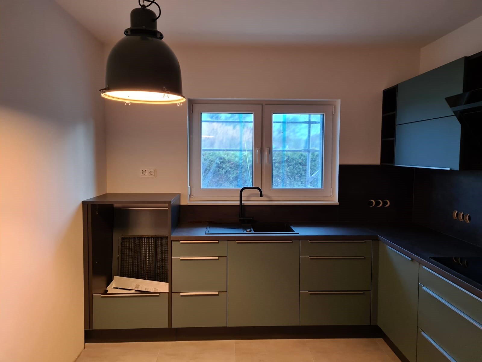 Küche mit grünen Schränken, schwarzer Arbeitsplatte, Spüle unter Fenster, Pendelleuchte links.