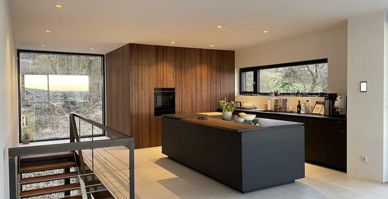 Moderne Küche mit dunkler Insel, Holzwand, Backofen, breite Fensterfronten und Treppengeländer