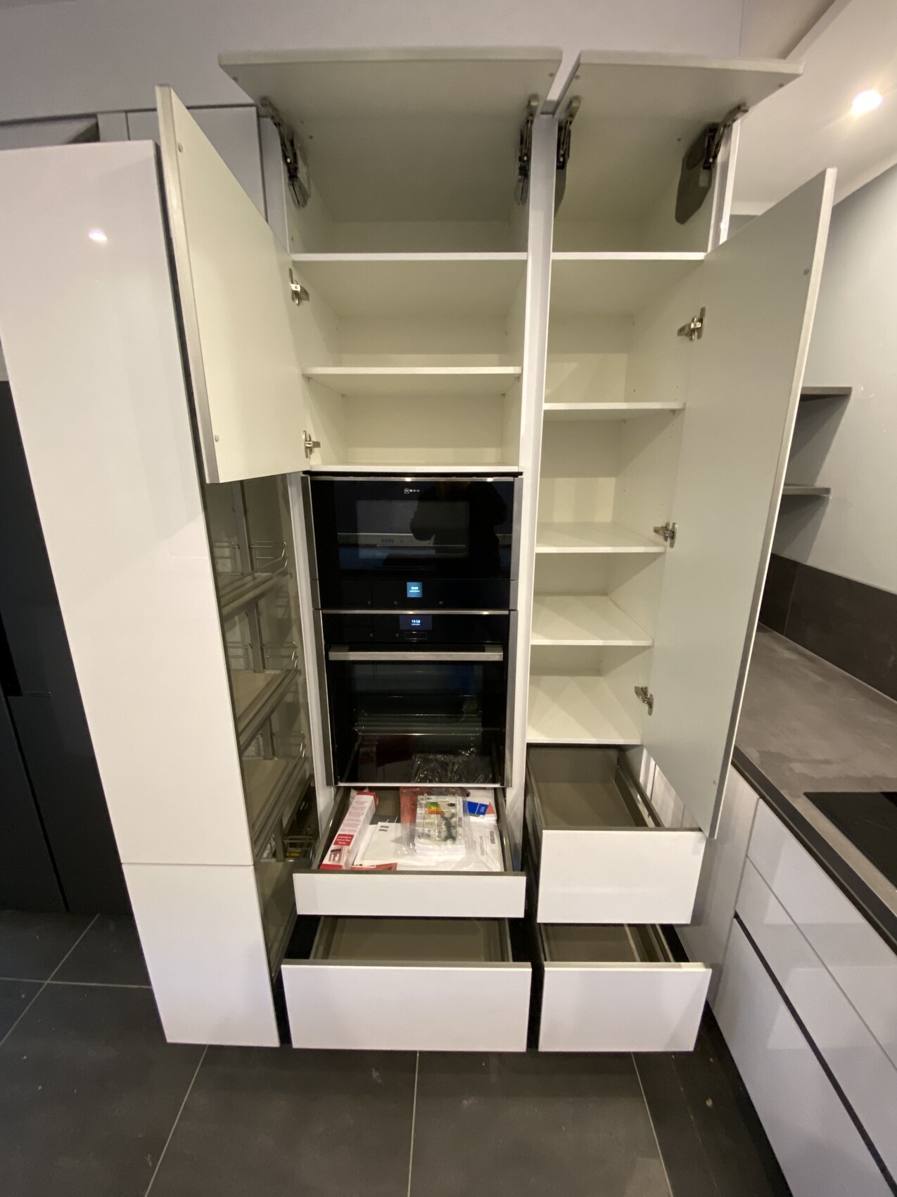 Moderne weiße Küche: zentrale schwarze Backöfen, offene Regale, unten Schubladen.