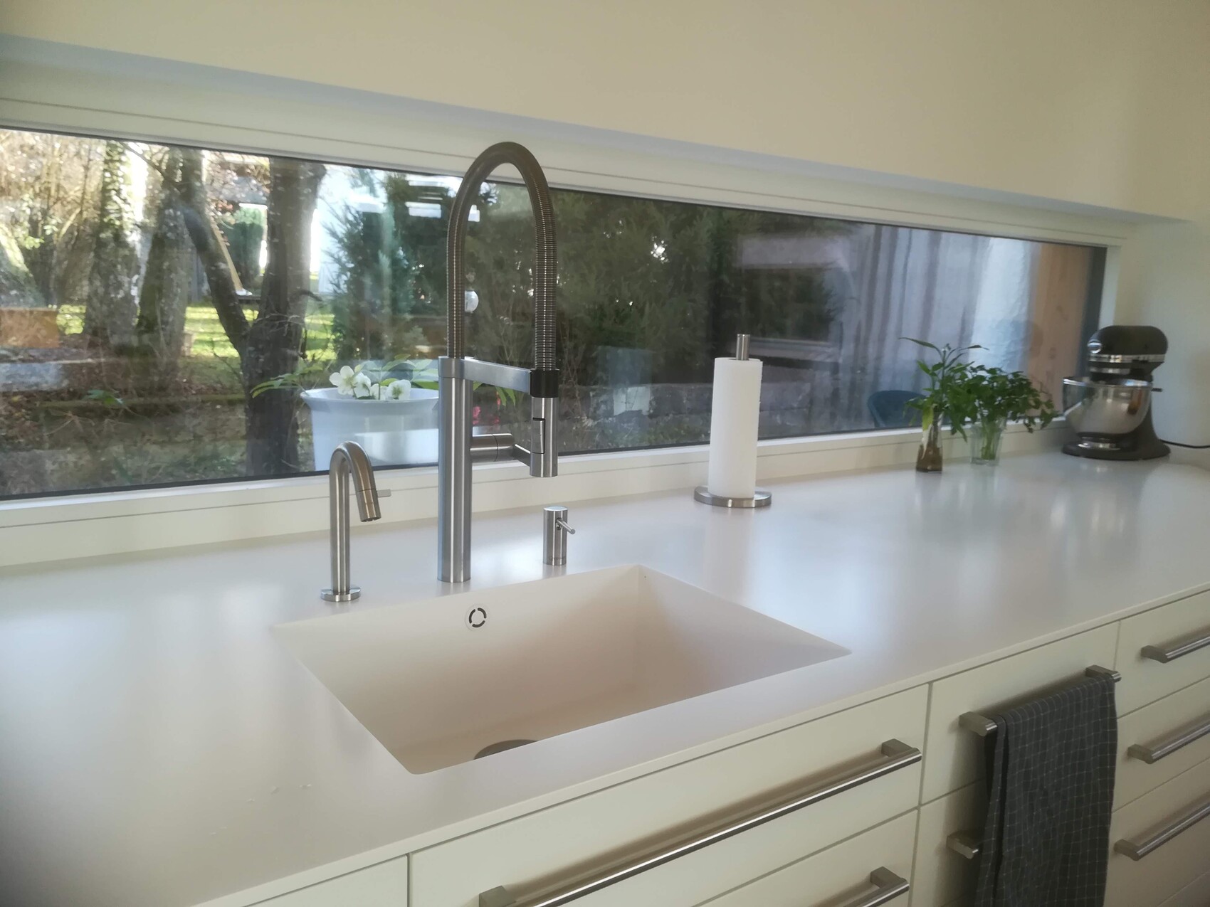 Moderne Küchenspüle mit Hochbogenarmatur, Papierrolle, Kräutertöpfchen und Standmixer am Fenster.