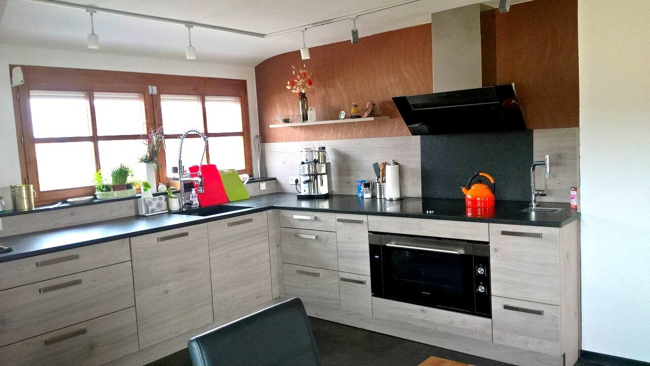 Moderne Küche mit grauen Holzschränken, schwarzer Arbeitsplatte, Backofen, Spüle und orange Kessel.