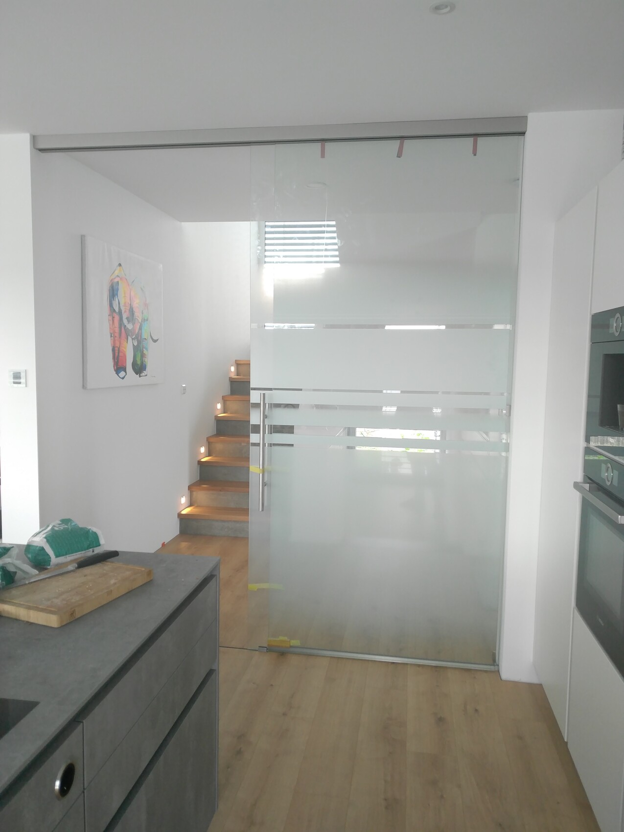 Moderne, offene Wohnküche mit Glas-Schiebetür, Holztreppe und bunter Elefanten-Wandkunst.