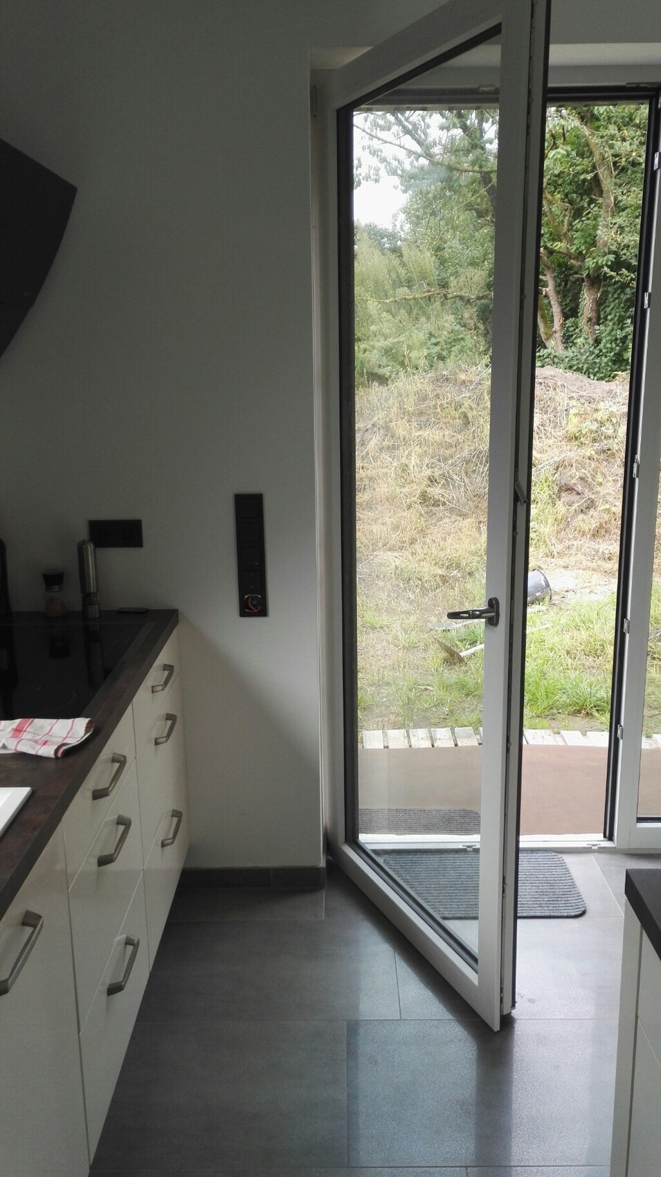 Moderne Küche mit weißen Schränken links; Glastür öffnet nach draußen zum Garten.
