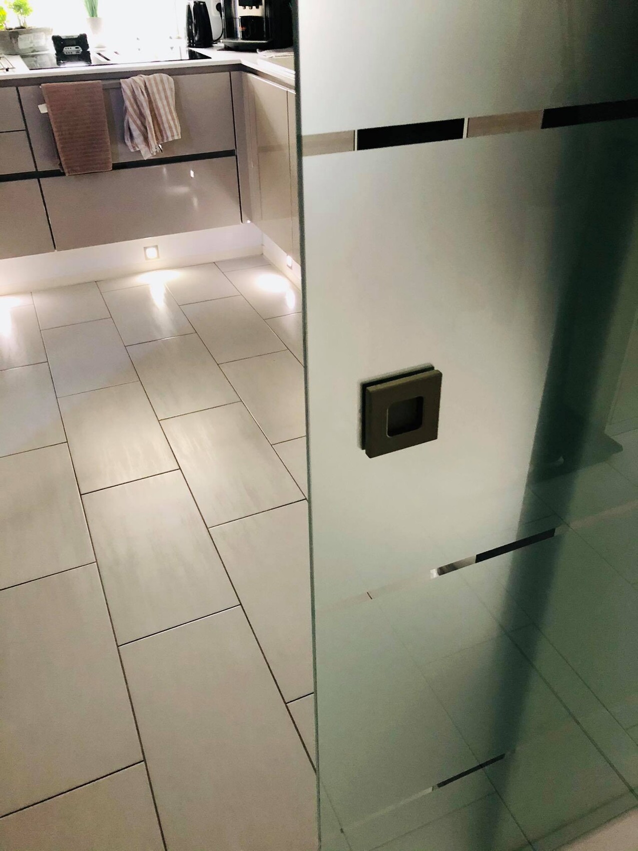 Moderne Küche mit grauen Hochglanz-Schränken, Fliesenboden und Glas-Trennwand im Vordergrund.