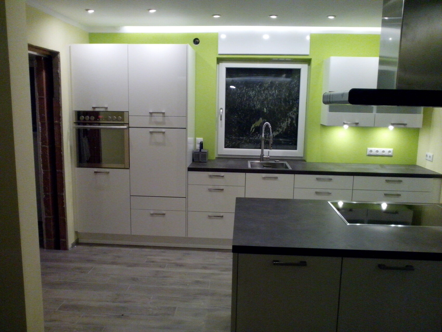 Moderne Küche mit weißen Schränken, Backofen, Spüle vor Fenster und grauen Arbeitsplatten.
