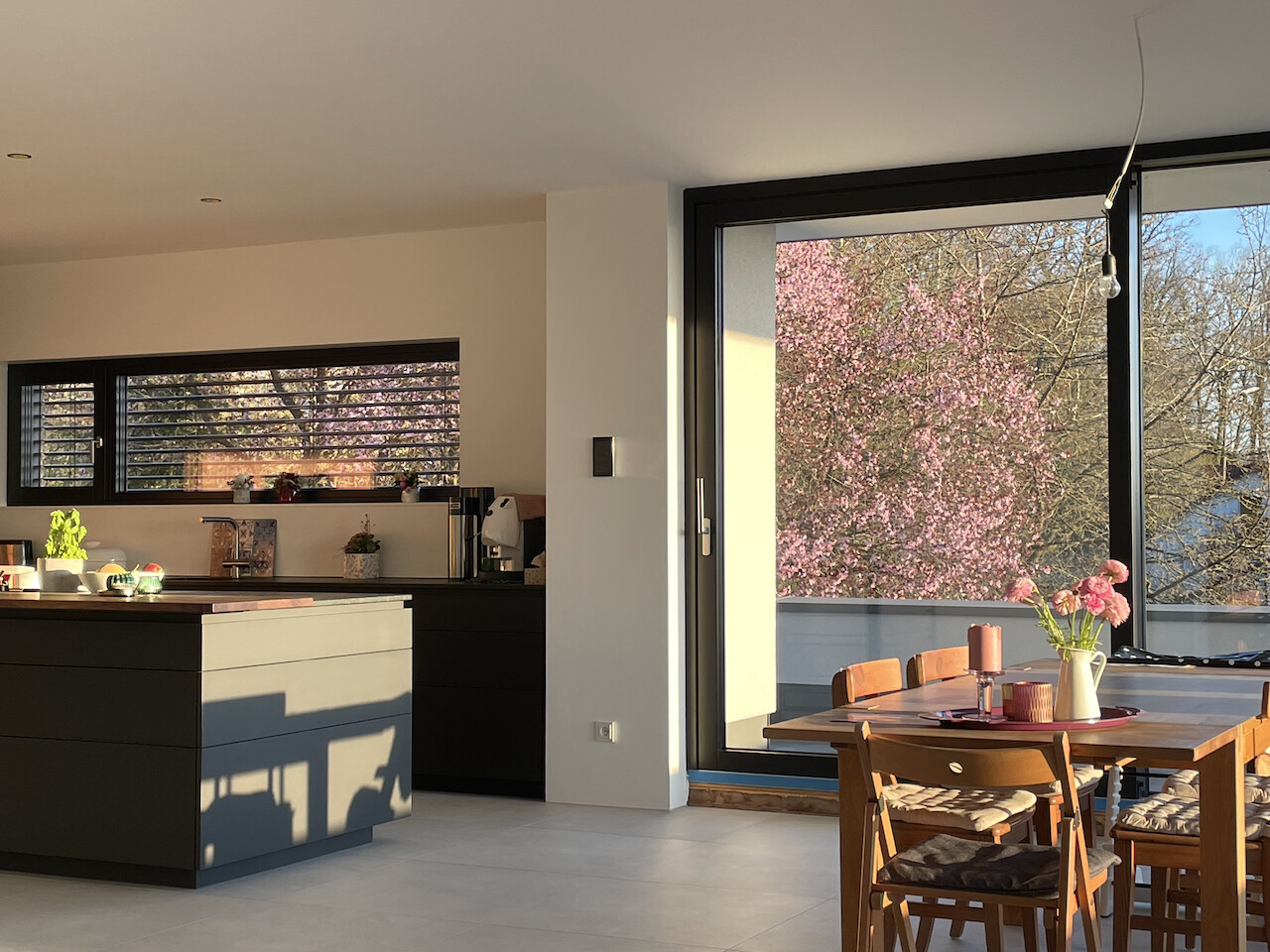 Moderne Küche mit Insel und Essbereich; grosse Glasfront zur Terrasse mit Kirschblüten.
