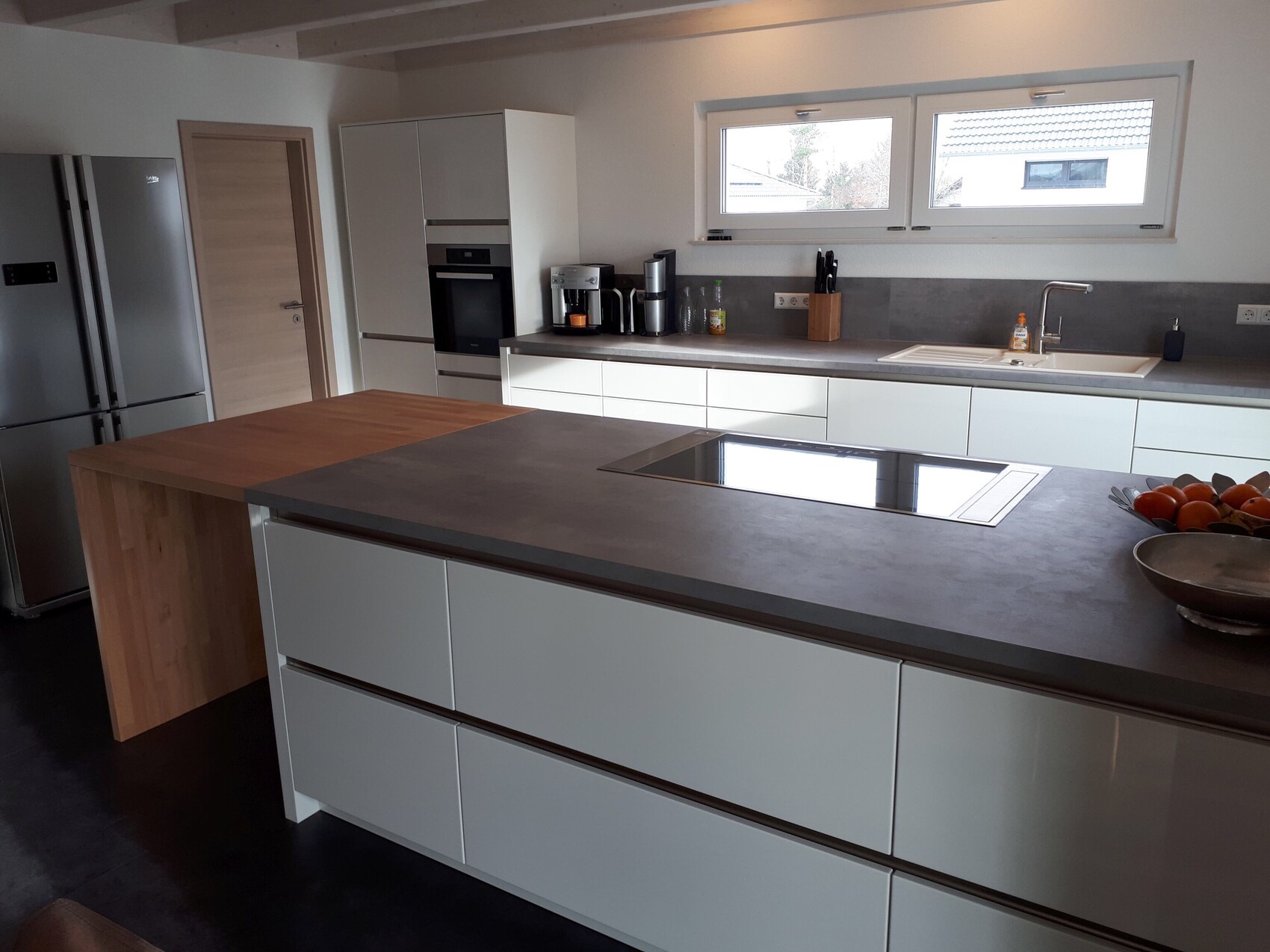 Moderne Küche mit großer Insel, Induktionskochfeld, Spüle, Backofen, Kühlschrank und Fenster.