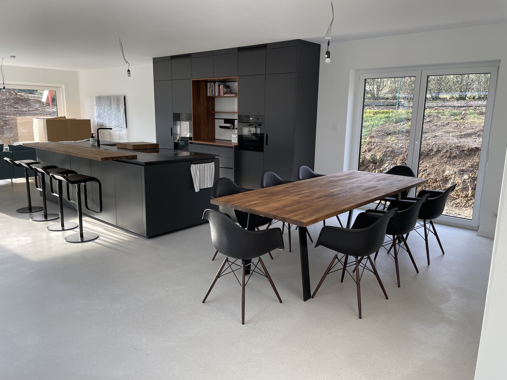 Moderne Küche mit dunkler Kücheninsel, Barhockern, Holz-Esstisch und großen Glastüren nach draußen.