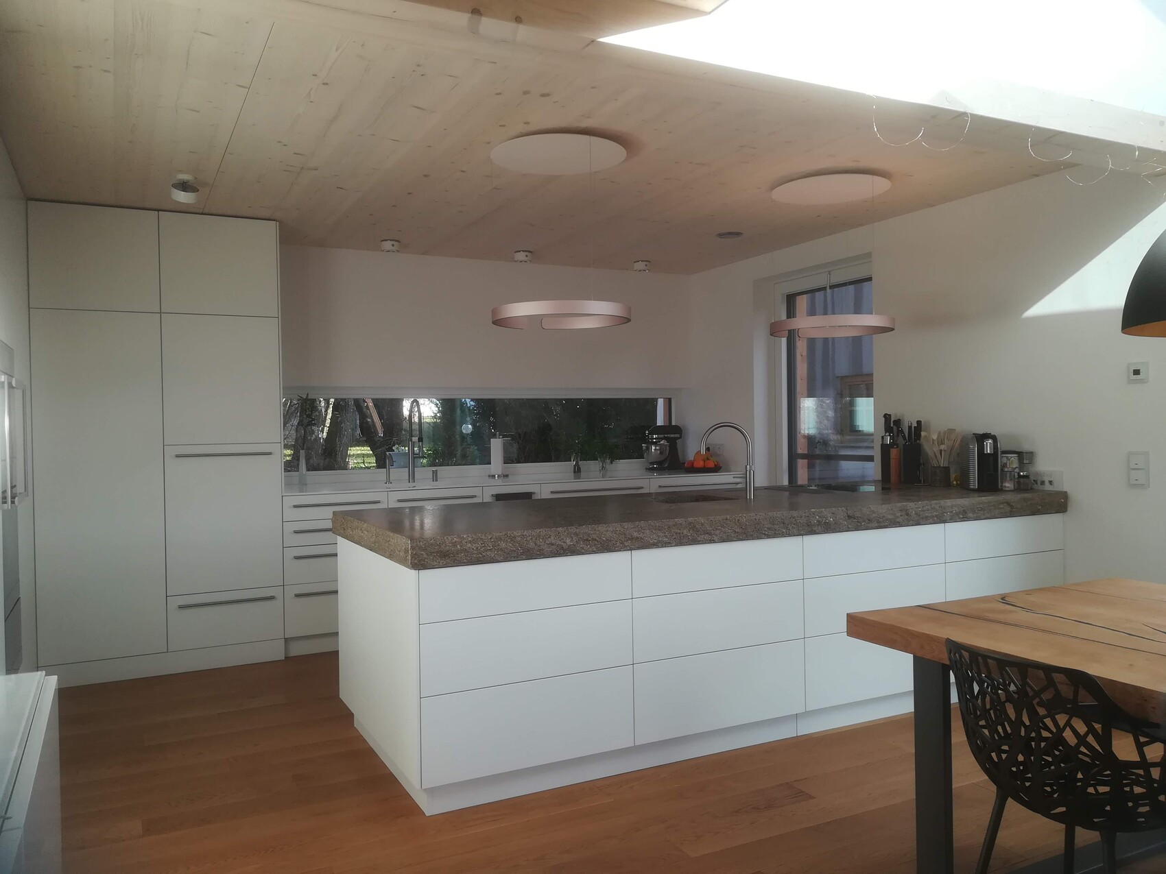 Moderne weiße Küche mit Insel, Holzfußboden, grauer Steinplatte und pinken Pendelleuchten.