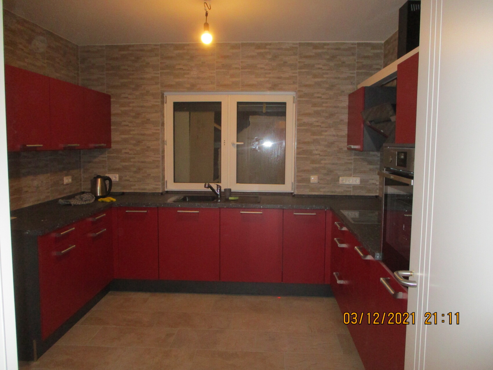 Küche mit roten Unterschränken, grauer Fliesenrückwand, Spüle unter Fenster, Backofen rechts.