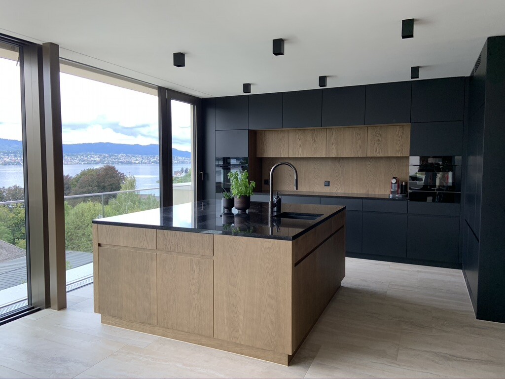 Moderne Küche mit dunklen Fronten, Eichenholz-Insel, Spüle und Panoramablick aufs Wasser.
