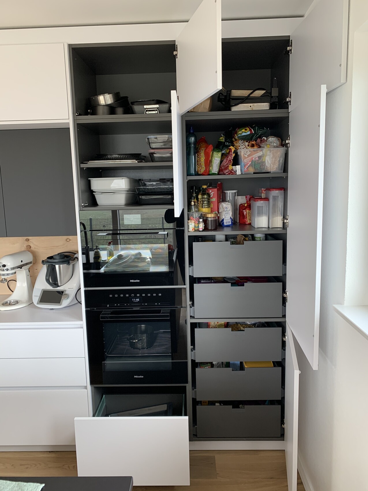 Offene Küchenschränke mit Miele-Backofen, Töpfen, Vorratsdosen und Schubladen voller Lebensmittel