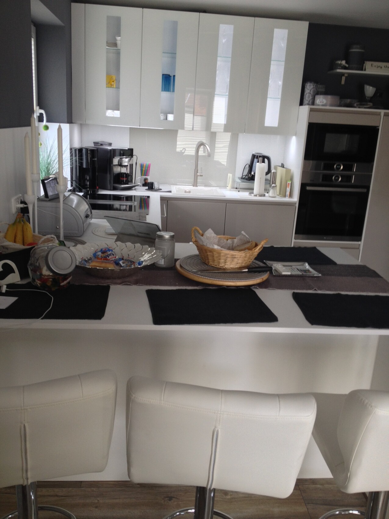 Moderne weiße Küche mit Insel, vier Barhockern, Obstkorb, Kaffeemaschine und Backofen.