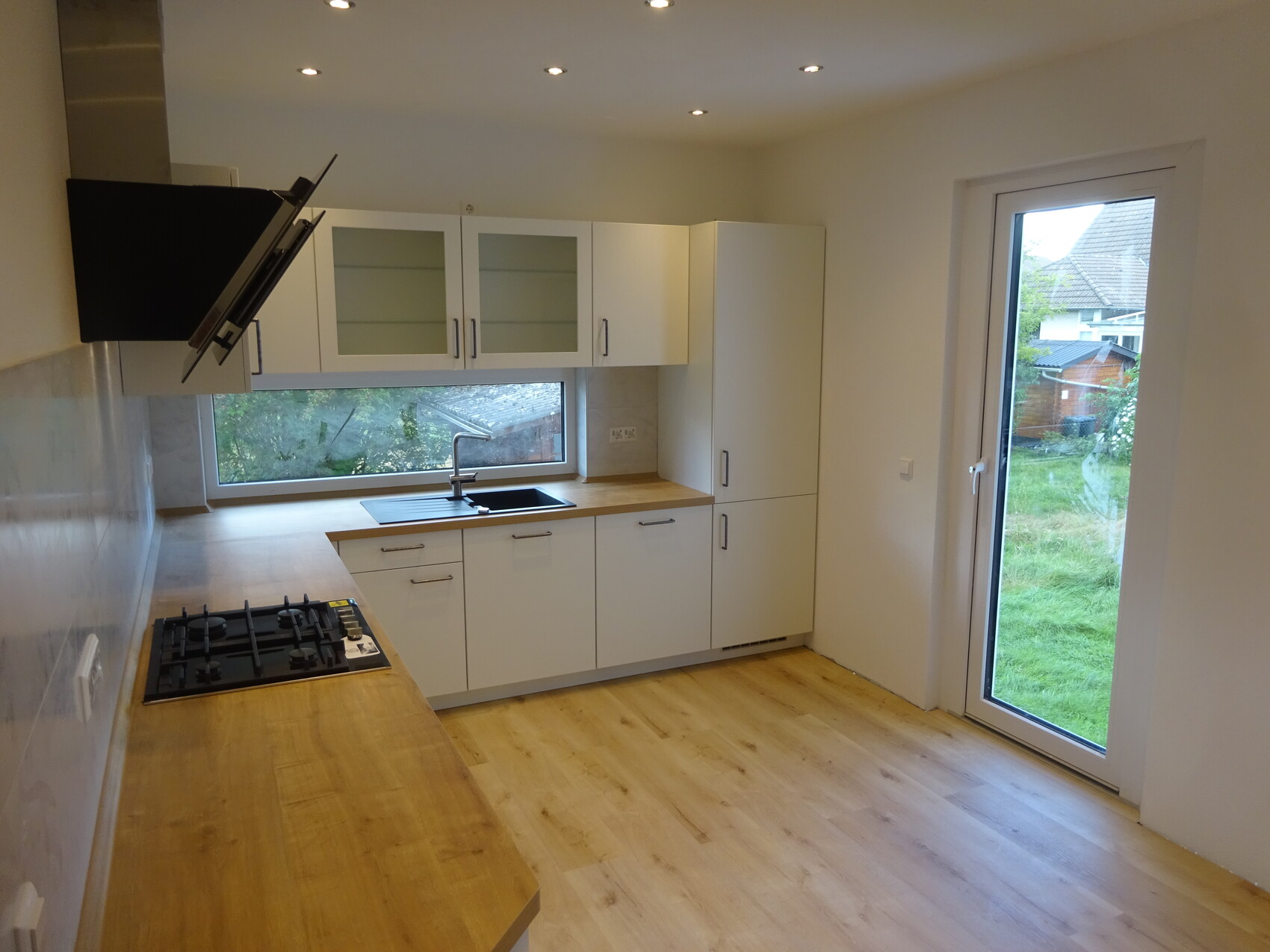 Moderne Küche: weiße Schränke, Holz-Arbeitsplatte, Gasherd links, Spüle unter Fenster, helle Räume.