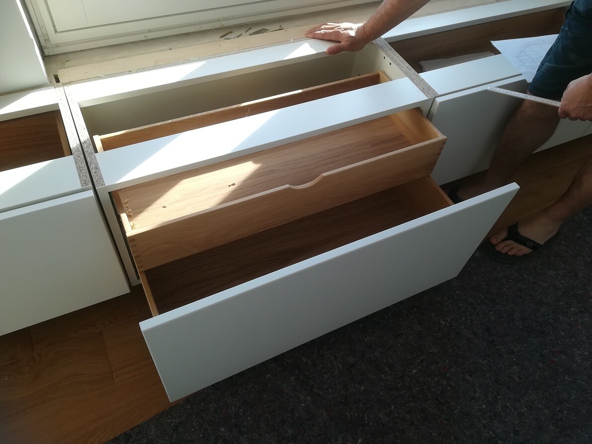 Person legt Maßband an weiße Küchenschubladen; mehrere Schubladen offen, Holzinnenraum sichtbar.
