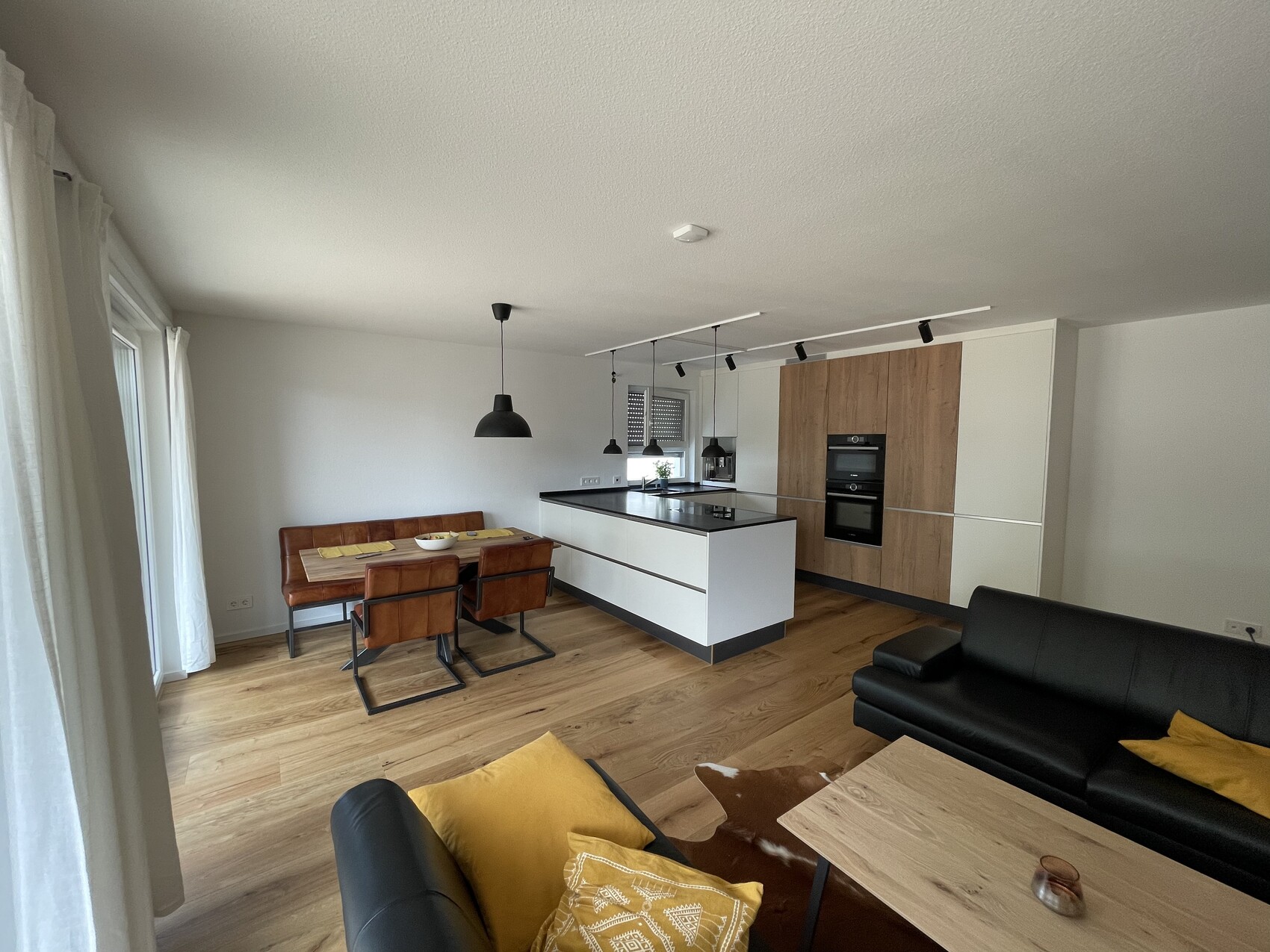 Offene Wohnküche mit Kochinsel, Esstisch, braune Stühle, schwarzes Sofa, gelbe Kissen, Holzboden.