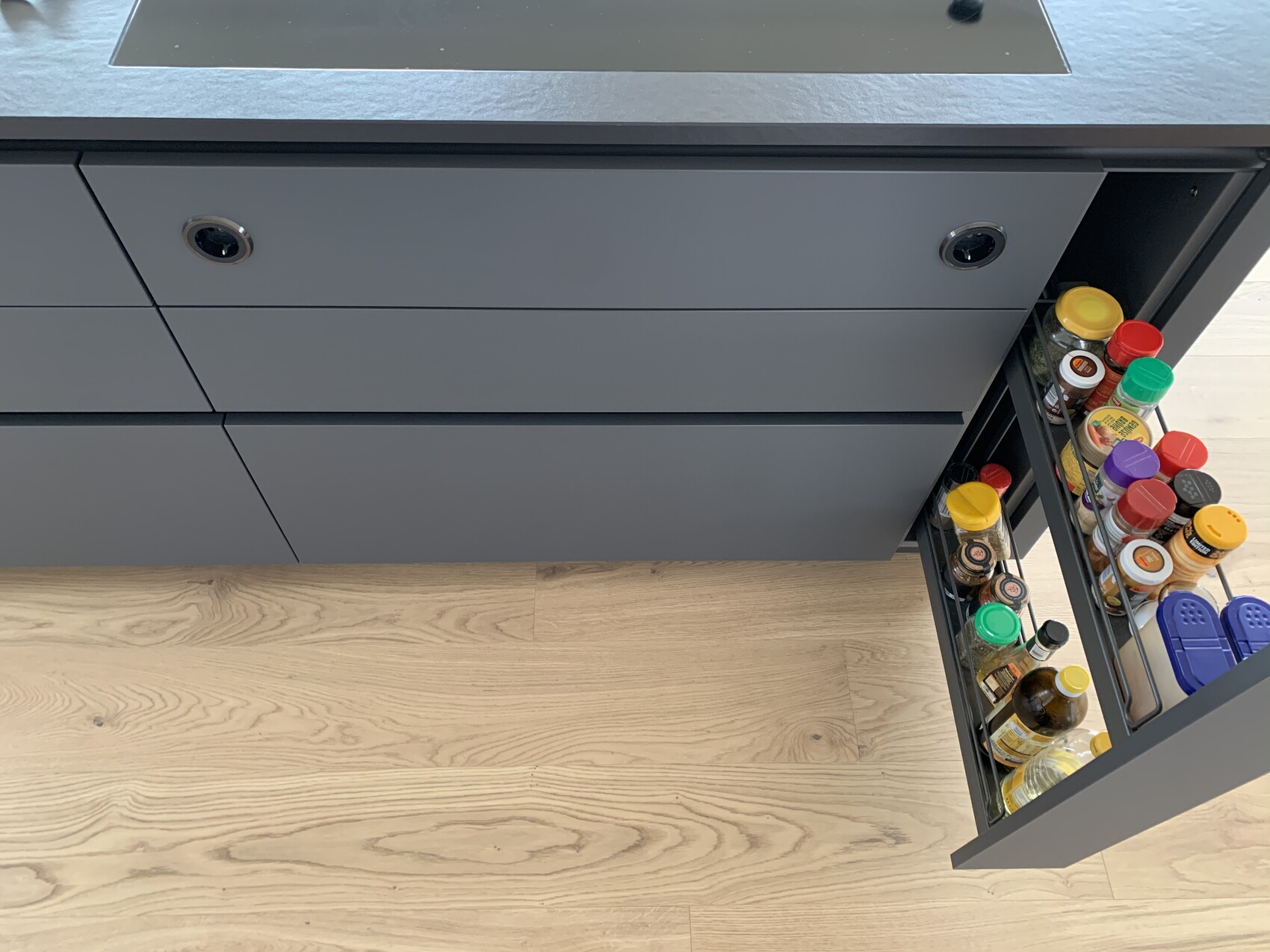 Grauer Küchenschrank mit ausziehbarer Gewürzschublade voller Flaschen und Gläser.