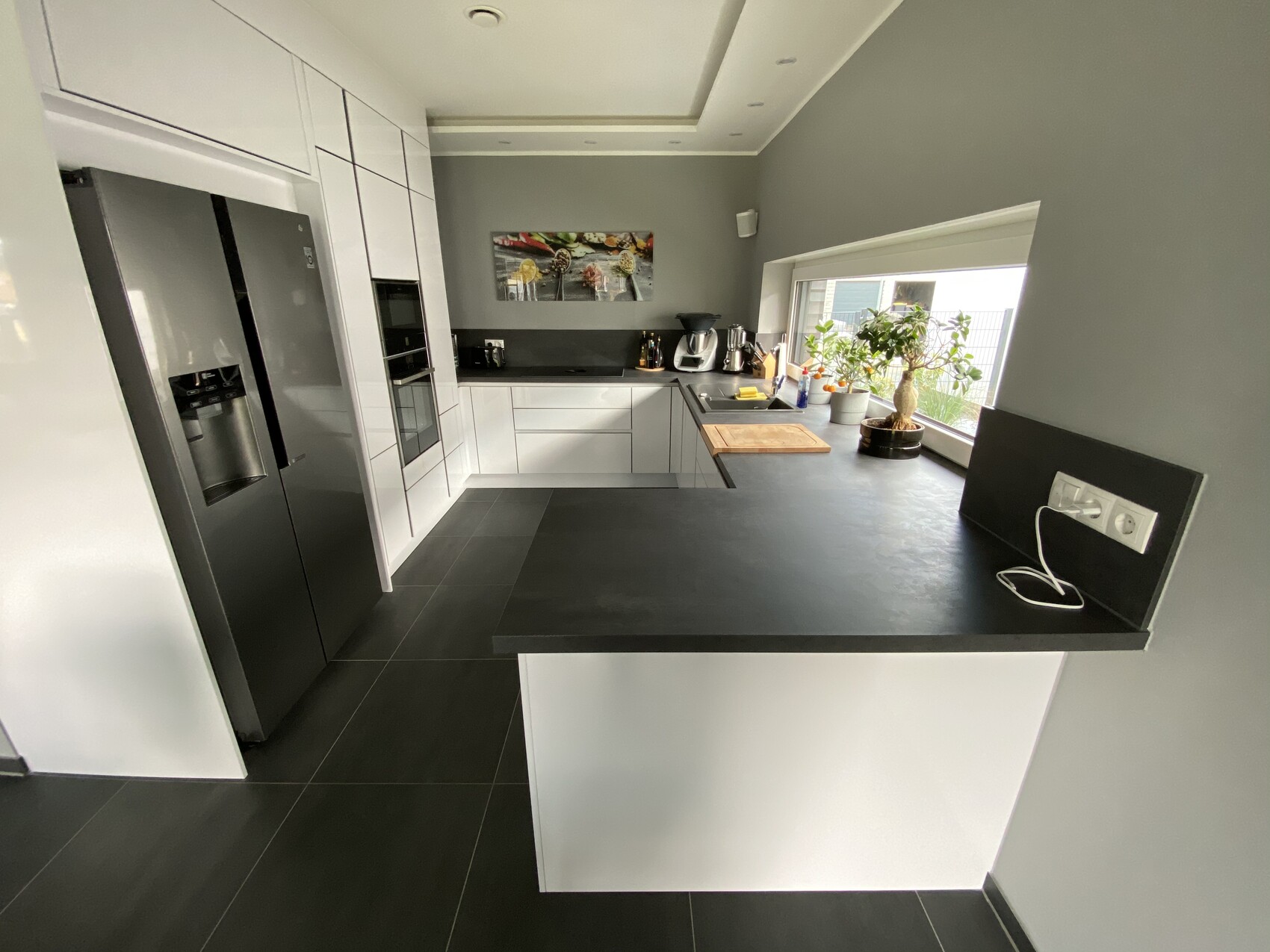 Moderne Küche mit weißen Schränken, schwarzen Arbeitsflächen, Kühlschrank, Ofen und Pflanze am Fenster.