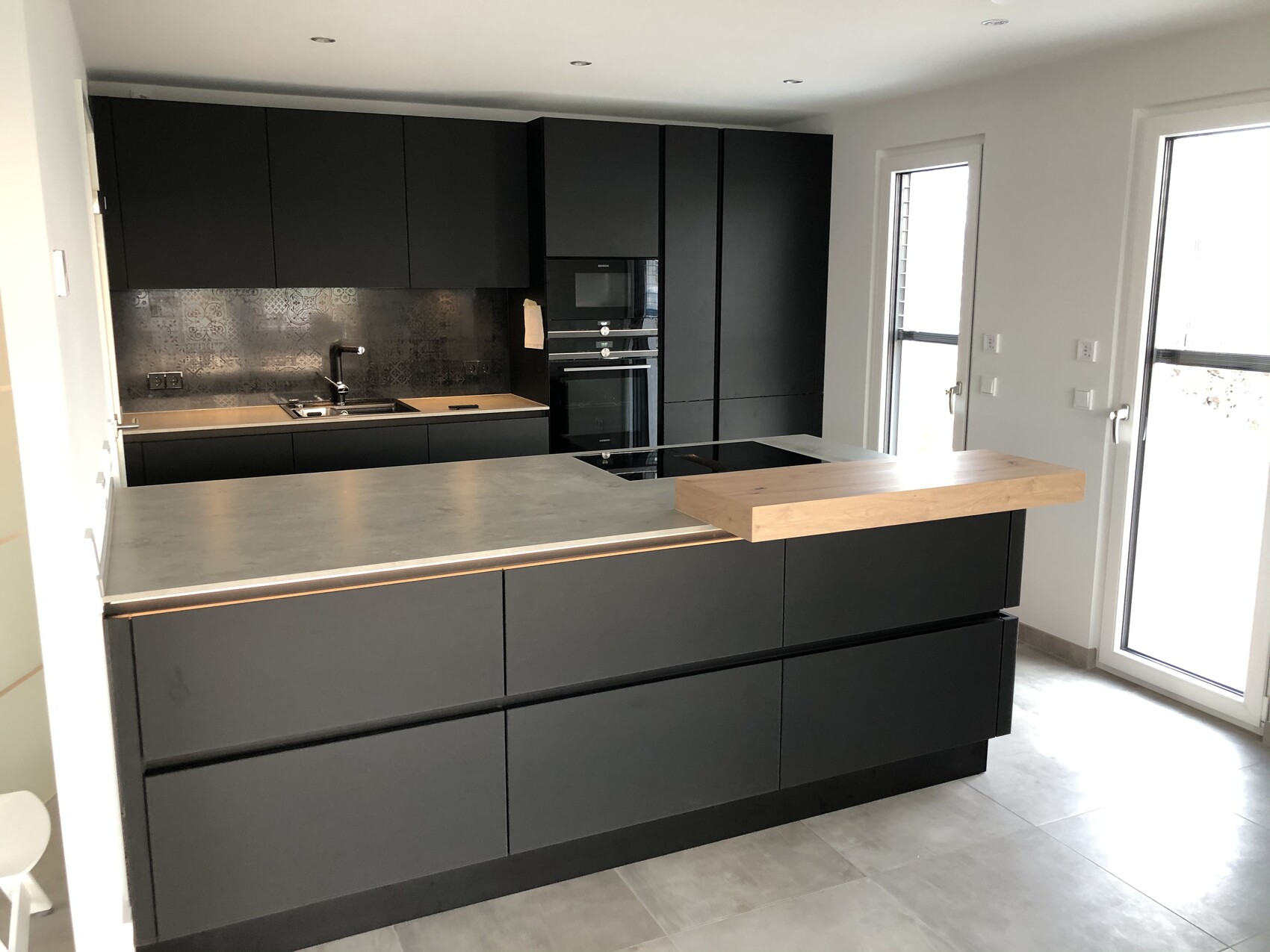 Moderne schwarze Küche mit Insel, Spüle und Backofen, viel Licht durch Fenster.