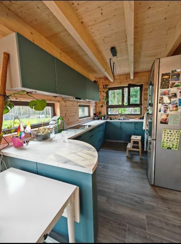 Küche in einem Holzhaus mit blauen Unterschränken, heller Arbeitsfläche, Fensterfront und Kühlschrank.