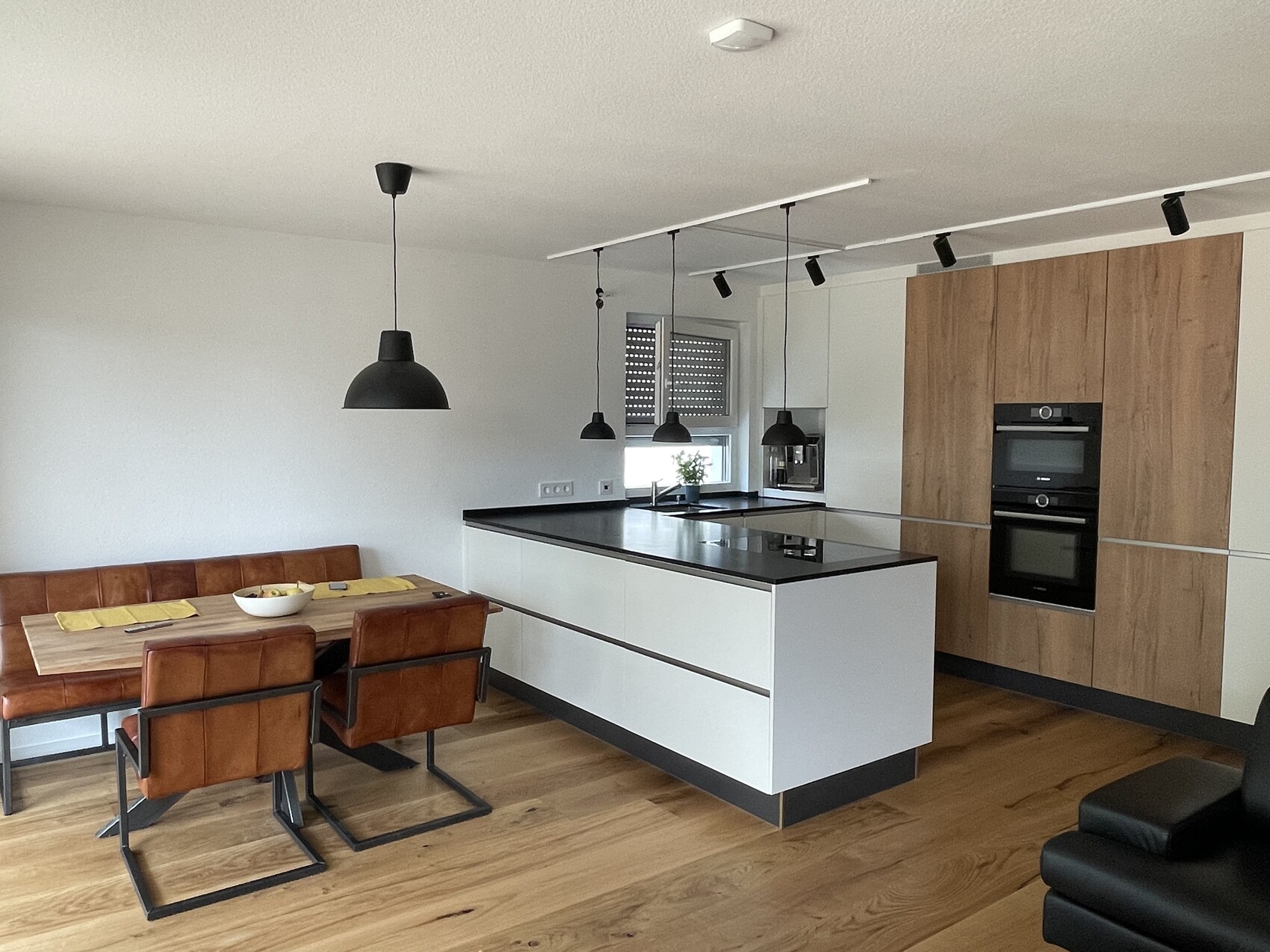 Moderne Küche mit Insel, schwarzen Arbeitsplatten, Holzschränken, Esstisch und dunklen Hängelampen.