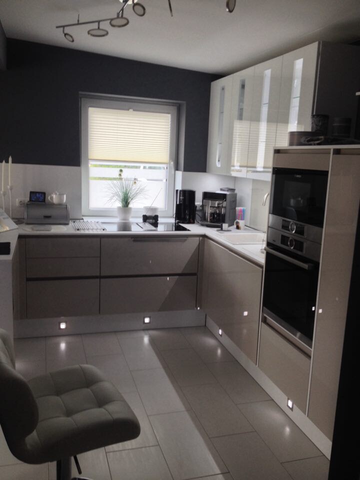 Moderne Küche mit weißen Hochglanz-Schränken, Einbauofen, Fenster mit Rollos, LED-Bodenlichtern.