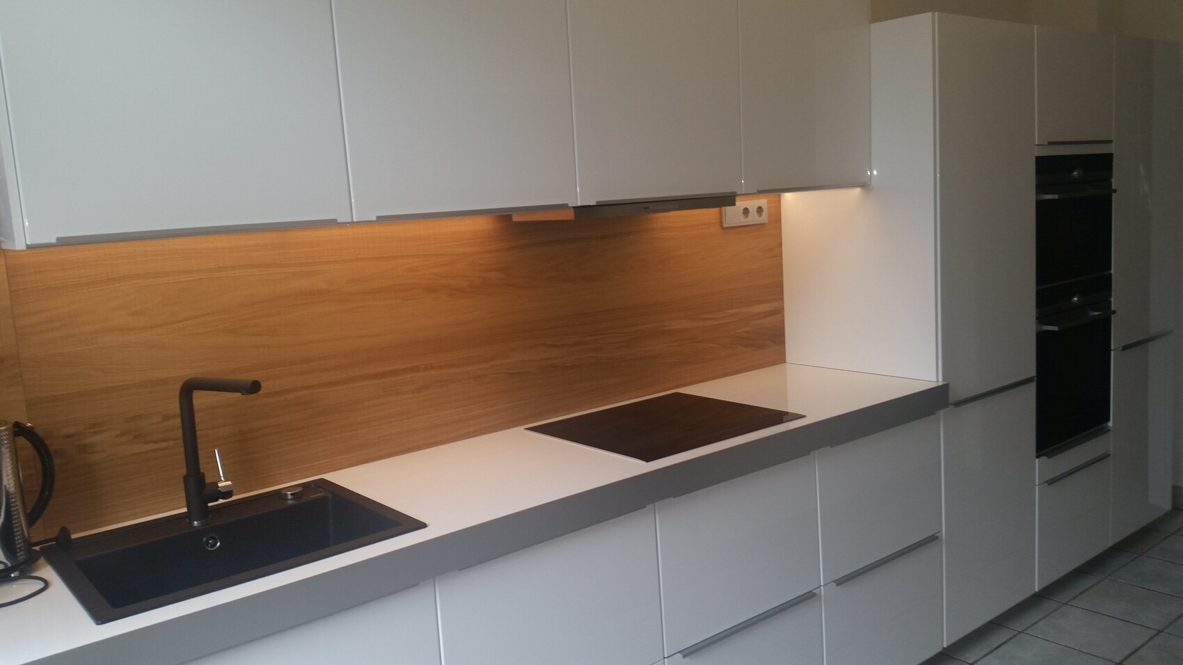 Moderne Küche mit weißen Hochglanzschränken, Holzrückwand, Spüle, Induktionskochfeld, Einbauofen.