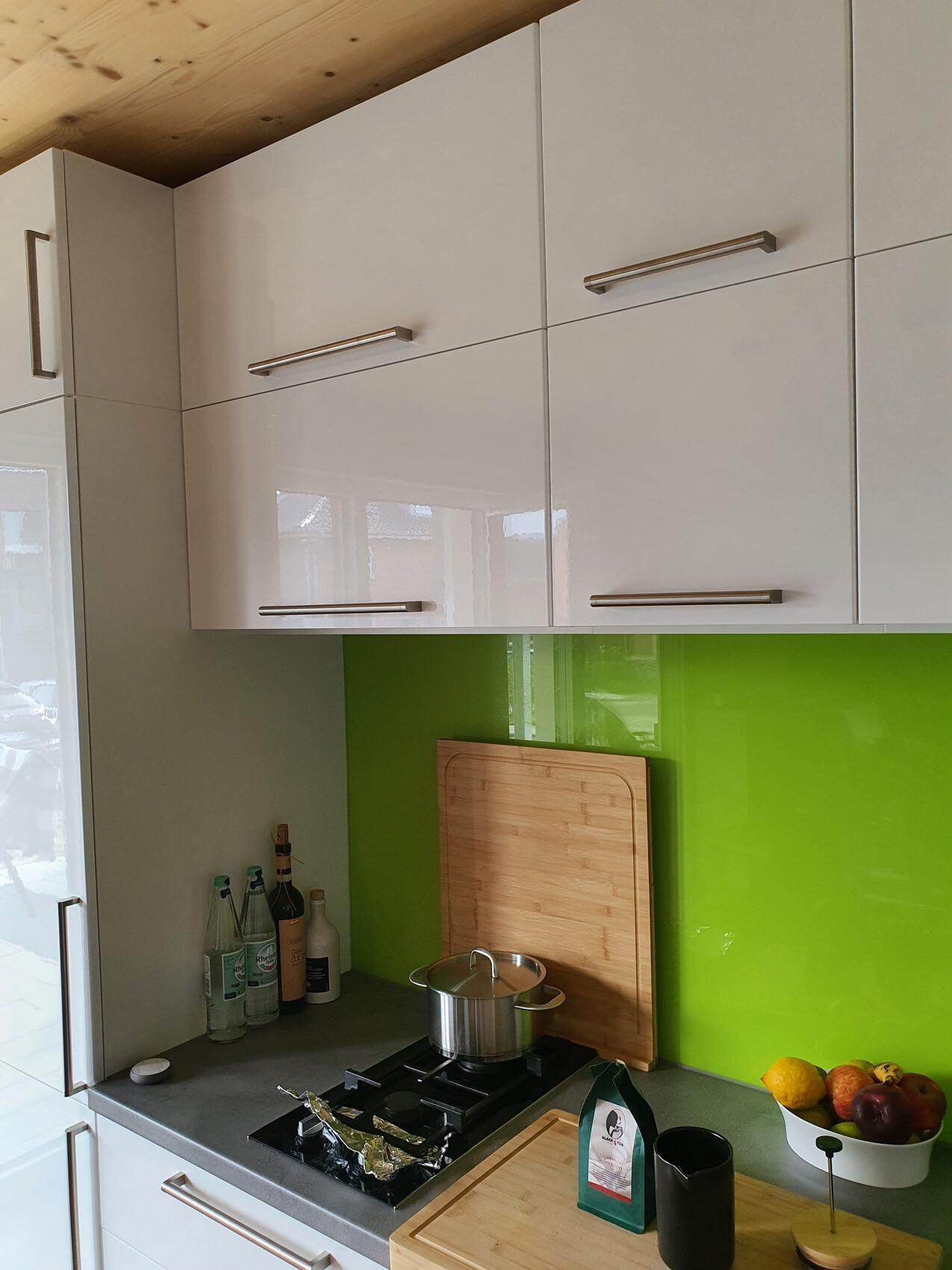 Küche mit weißen Hochglanz-Schränken, grüne Rückwand, Topf auf Gasherd, Obstschale.