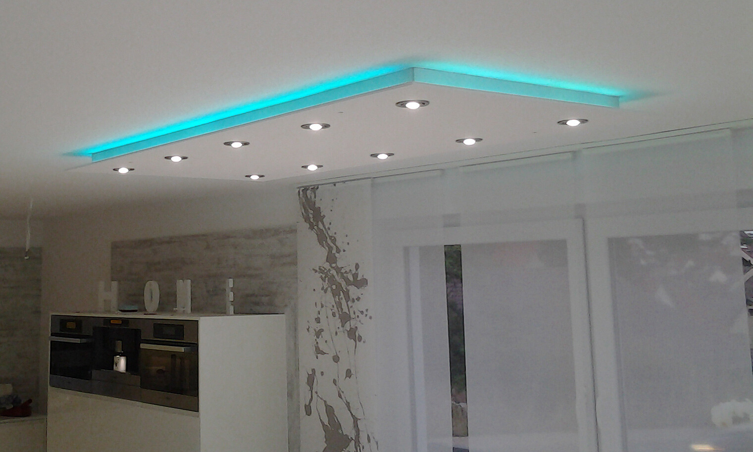 Moderne weiße Küche mit Deckenpaneel, cyan LED-Leiste und Spots; Einbau-Backöfen links.