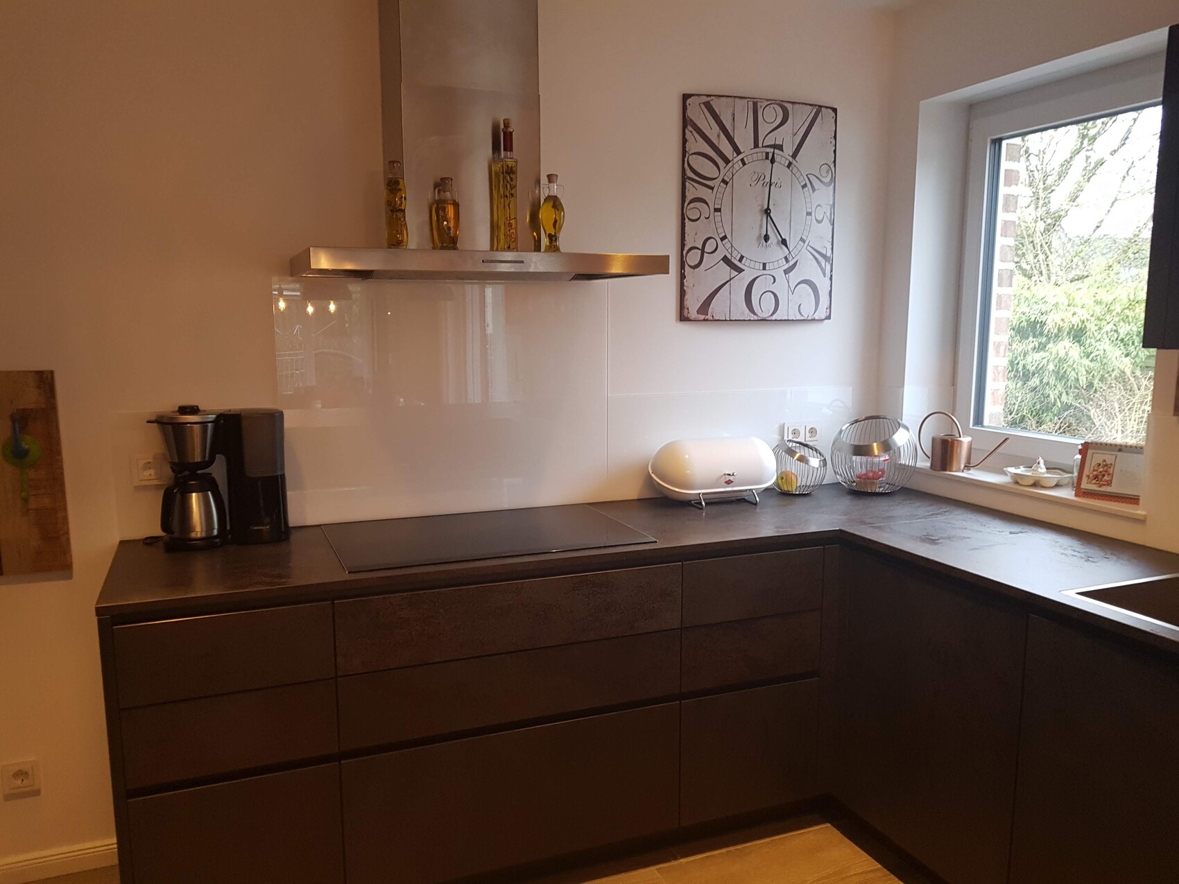 Moderne Küche mit dunklen Schränken, Dunstabzug, Kaffeeautomat, weißem Toaster, Obstkörbe und Wanduhr.