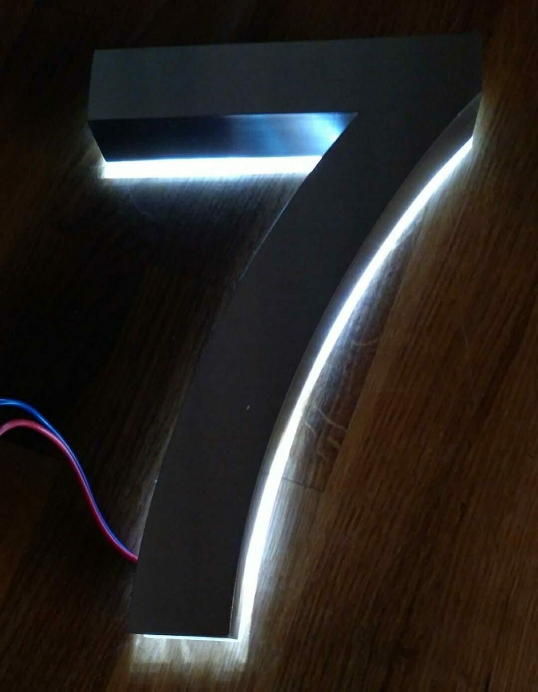 Großes schwarzes 7-Symbol mit weißen LED-Lichtkanten auf Holzboden, Kabel sichtbar.