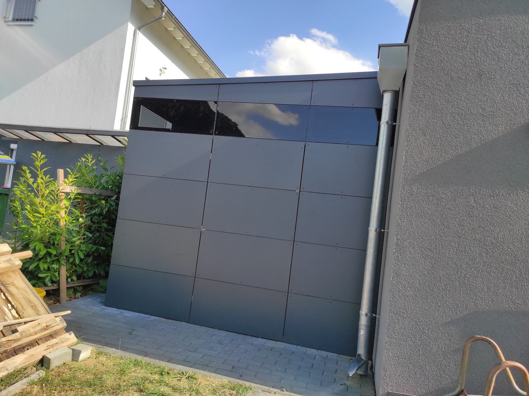 Außenansicht einer grauen Paneelwand mit reflektierenden Oberflächen, Dachrinne und Holzstapel.