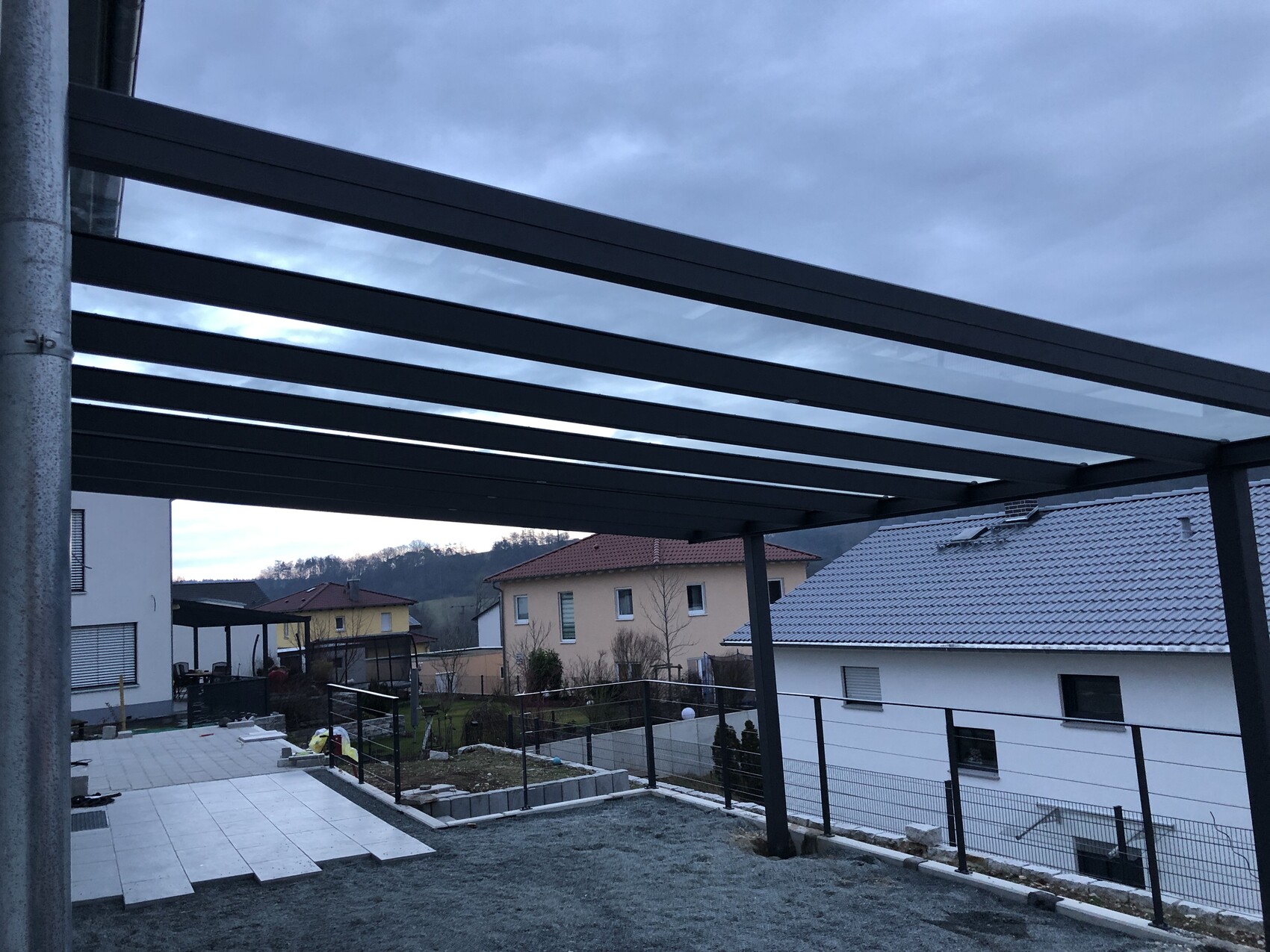Stahlträger-Pergola im Vorgarten, Terrasse und Garten, Nachbarhäuser mit Ziegeldächern.