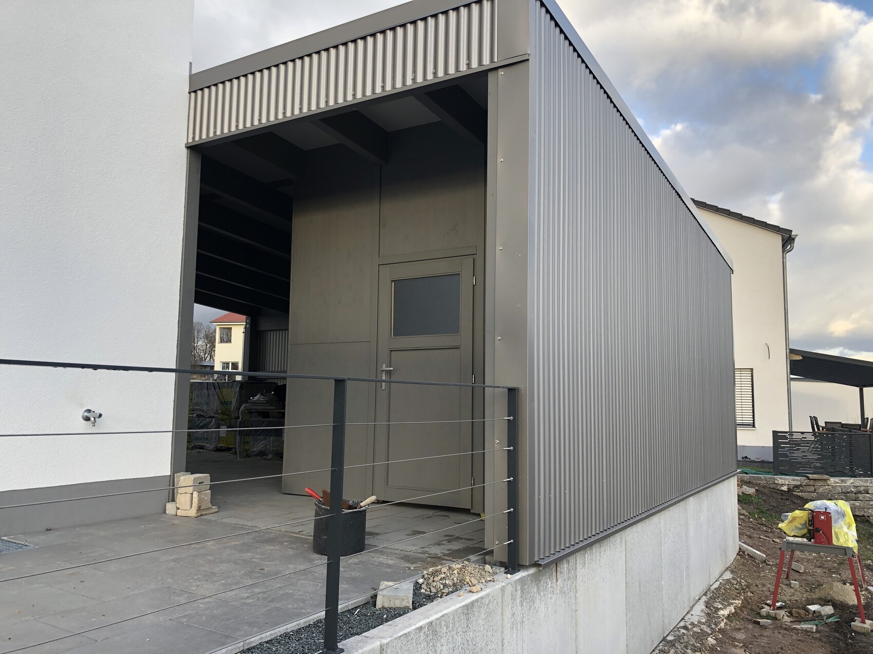 Moderner Anbau aus grauer Metallverkleidung mit Tür, Geländer und Bauwerkzeugen vor einer Baustelle.