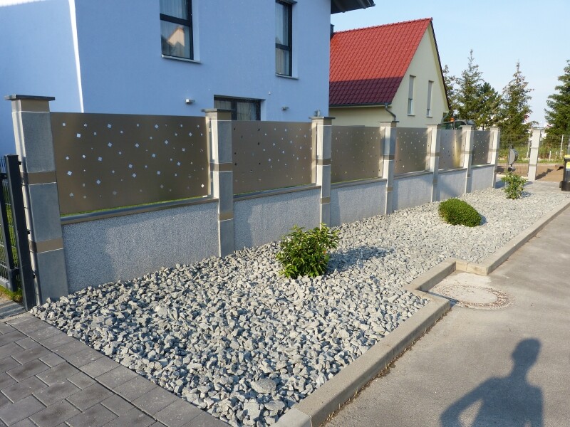 Moderner Metallzaun mit Lochblechen vor grauer Mauer, Kies und Sträucher.