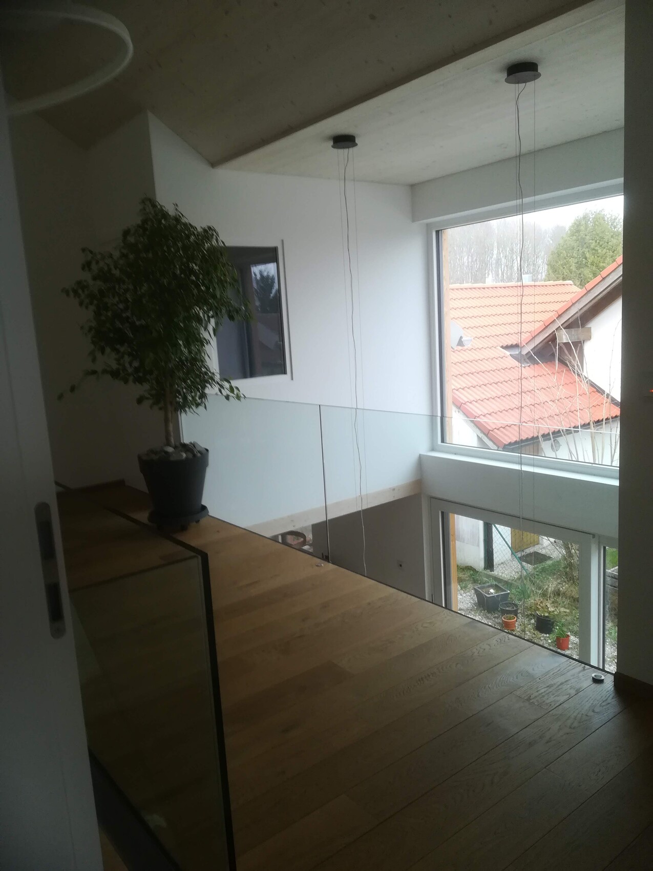 Loftartiger Innenraum mit Holzboden, Glasgeländer und Pflanze; Blick auf rotes Dach durch Fenster.