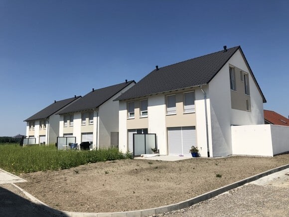 Moderne weiße Reihenhäuser mit Garagenfronten, davor unbefestigte Freifläche und blauer Himmel.