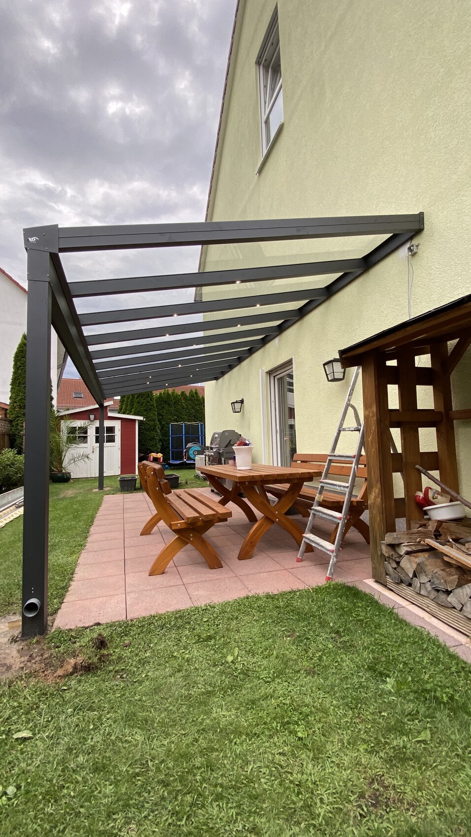 Garten mit Metallpergola über Terrasse, Holztisch mit Bänken, Leiter und Brennholzstapel.