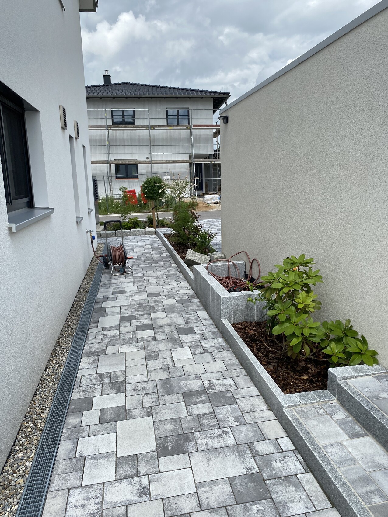Gepflasterter Weg zwischen weißen Hauswänden, Gartenbeet und Baugerüst im Hintergrund.