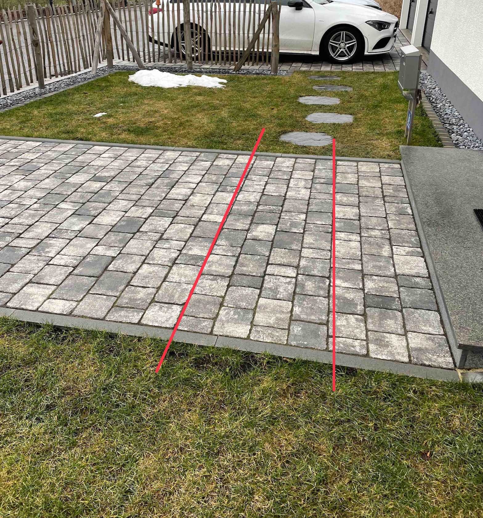 Gepflasterte Zufahrt aus grauen Pflastersteinen, Grasstreifen, weißes Auto hinter Holzzaun.