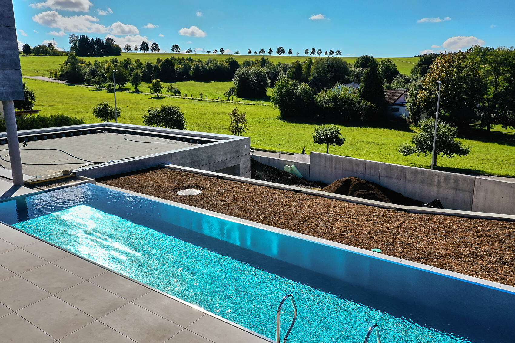 Moderne Terrasse mit Pool im Vordergrund, Baufläche und grüne Wiesen dahinter.