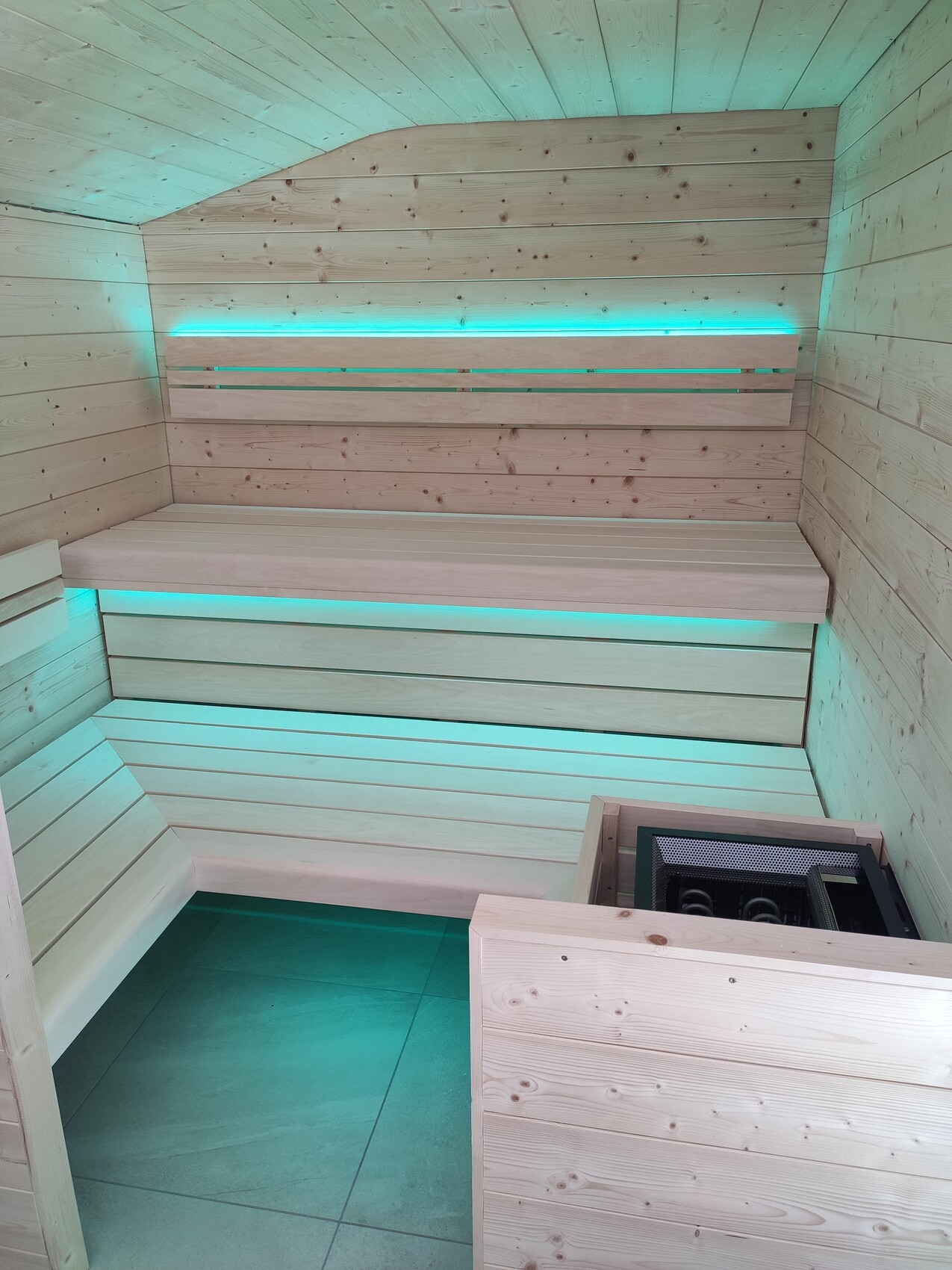 Holzsauna mit zwei Bänken, türkisfarbene Beleuchtung und Heizofen in der Ecke.