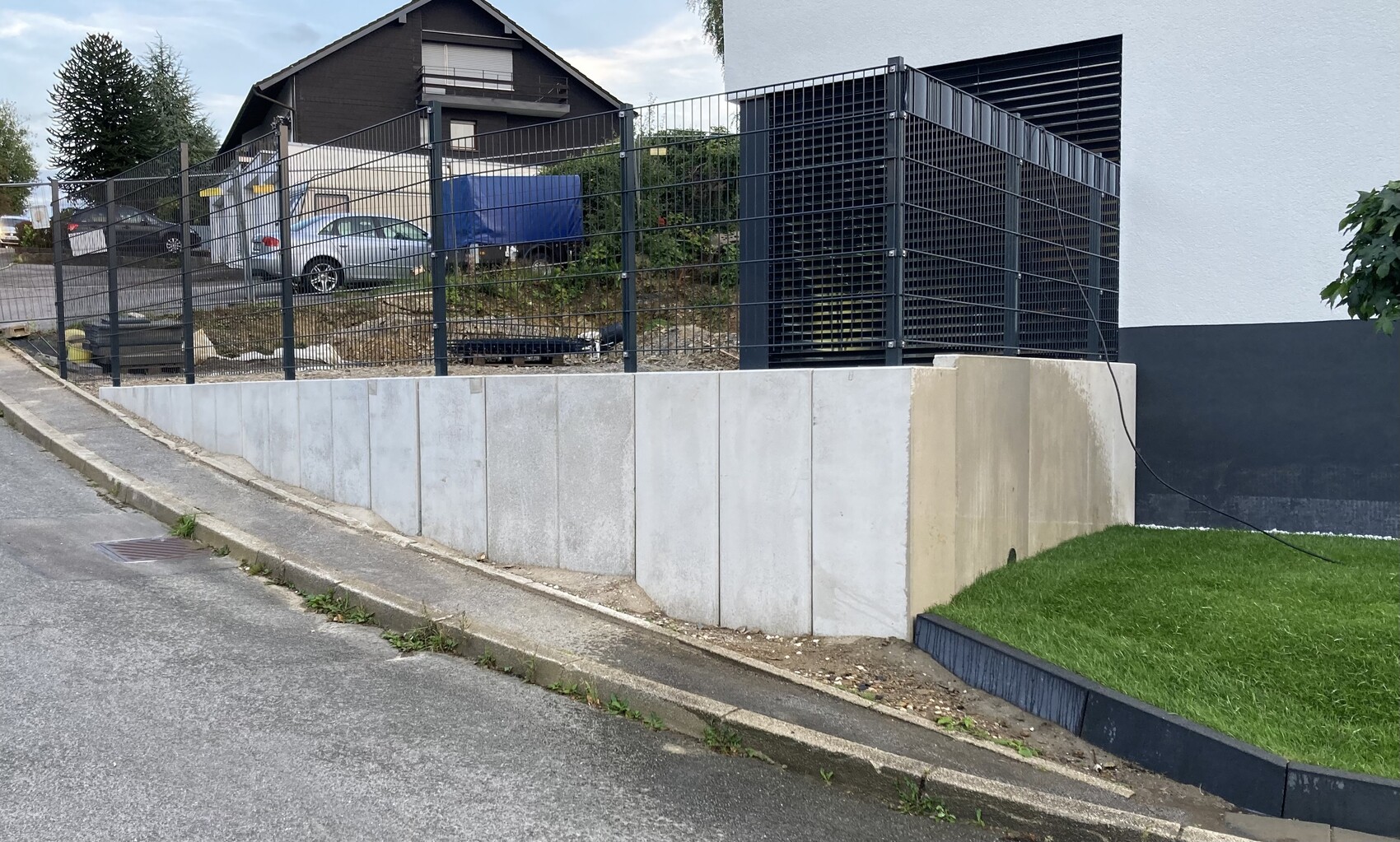 Betonplattenmauer mit schwarzem Metallzaun; dahinter parkende Autos und eine große Lüftungsanlage.