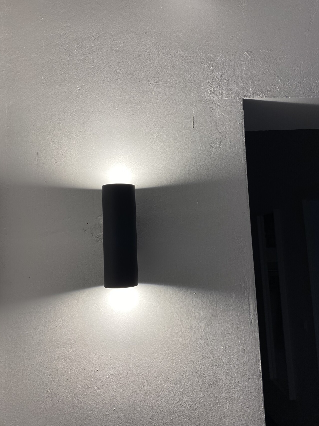 Schwarze, zylindrische Wandleuchte an heller Wand; Licht strahlt oben und unten.